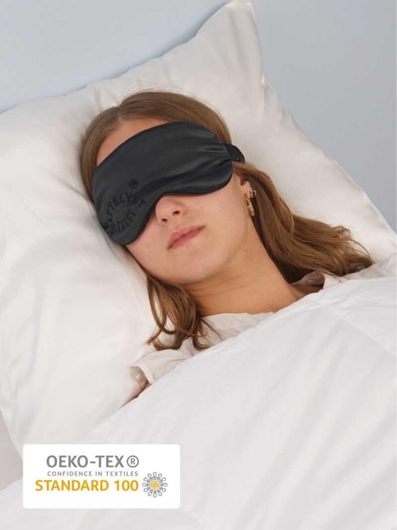 Junge Frau schläft mit schwarzer Schlafmaske aus Seide auf weißem Kissen und weißer Bettdecke, mit OEKO-TEX Standard 100 Label