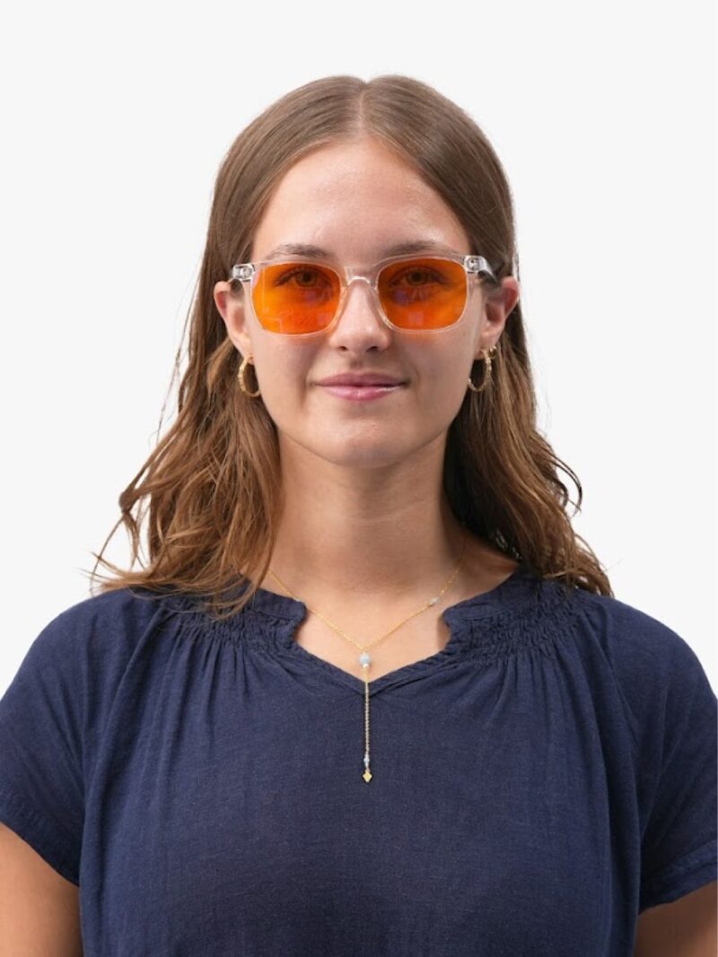 Junge Frau mit transparentem Gestell und orangefarbener Sonnenbrille, dunkelblaue Bluse, goldene Creolen und Kette mit Anhänger