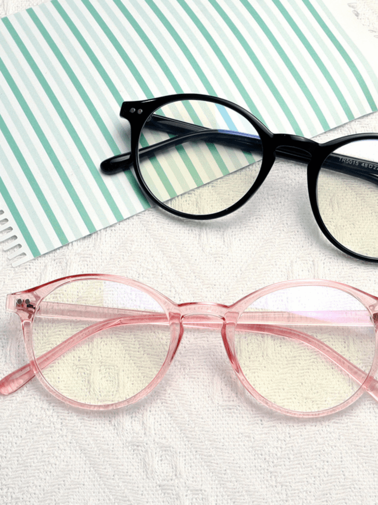 Schwarze runde Brille und transparent-rosa runde Brille auf weißem Strickuntergrund neben grün gestreiftem Papier
