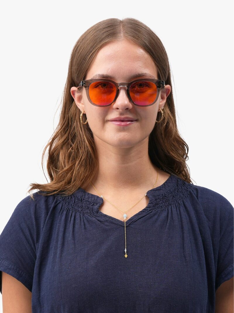 Frau mit grauer runder Brille mit orange verspiegelten Gläsern, blauer Bluse und goldenem Schmuck