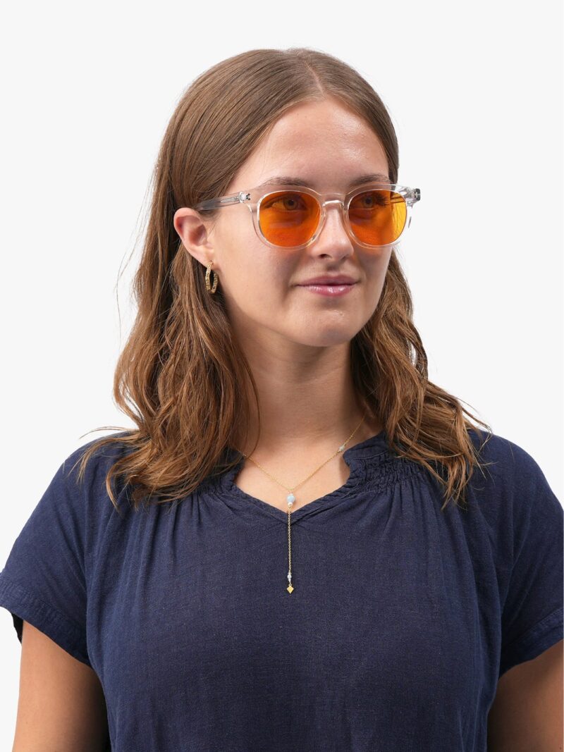 Junge Frau mit transparenter Brille mit orange getönten Gläsern, marineblauem T-Shirt, goldenem Anhänger und schulterlangem braunem Haar