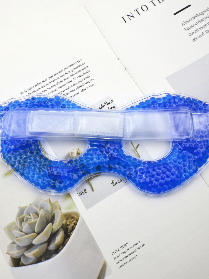 Blaue Gel-Augenmaske mit Kühlperlen und verstellbarem Klettband, abgelegt auf einer aufgeschlagenen Zeitschriftenseite
