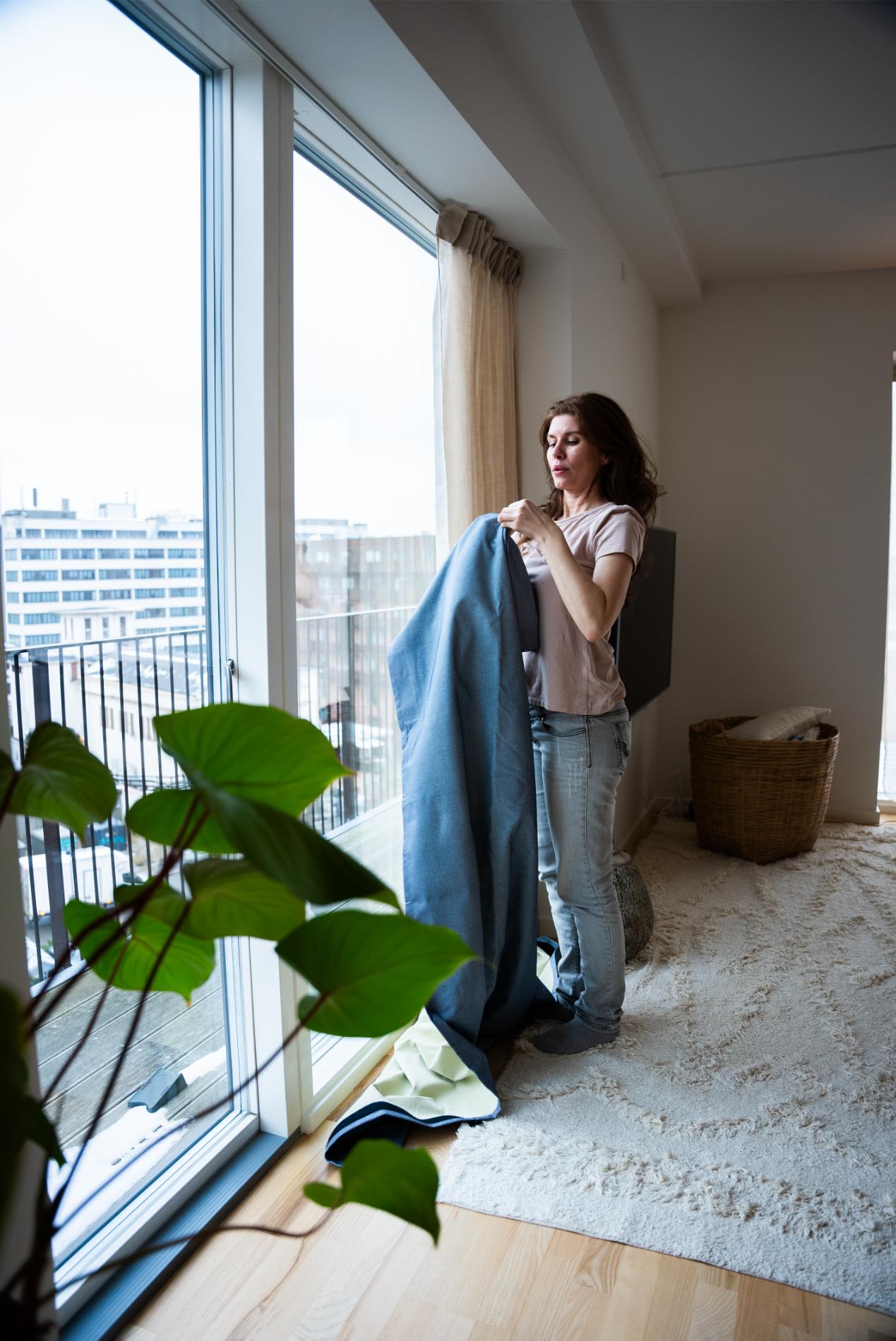 Frau richtet blauen Vorhang am großen Fenster einer Stadtwohnung, Pflanze im Vordergrund und Wäschekorb daneben