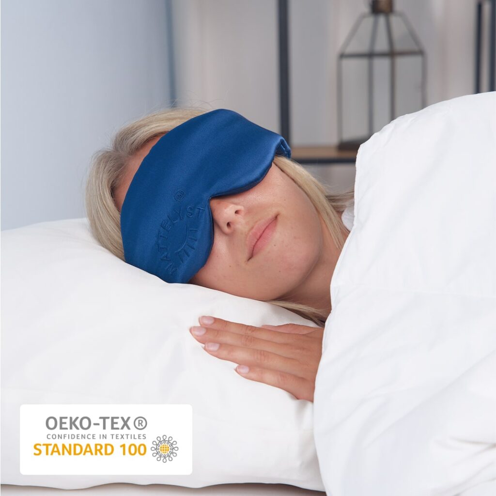 Schlafende Frau mit dunkelblauer Augenmaske auf weißem Kissen, Hand am Bett, OEKO-TEX STANDARD 100 Siegel sichtbar