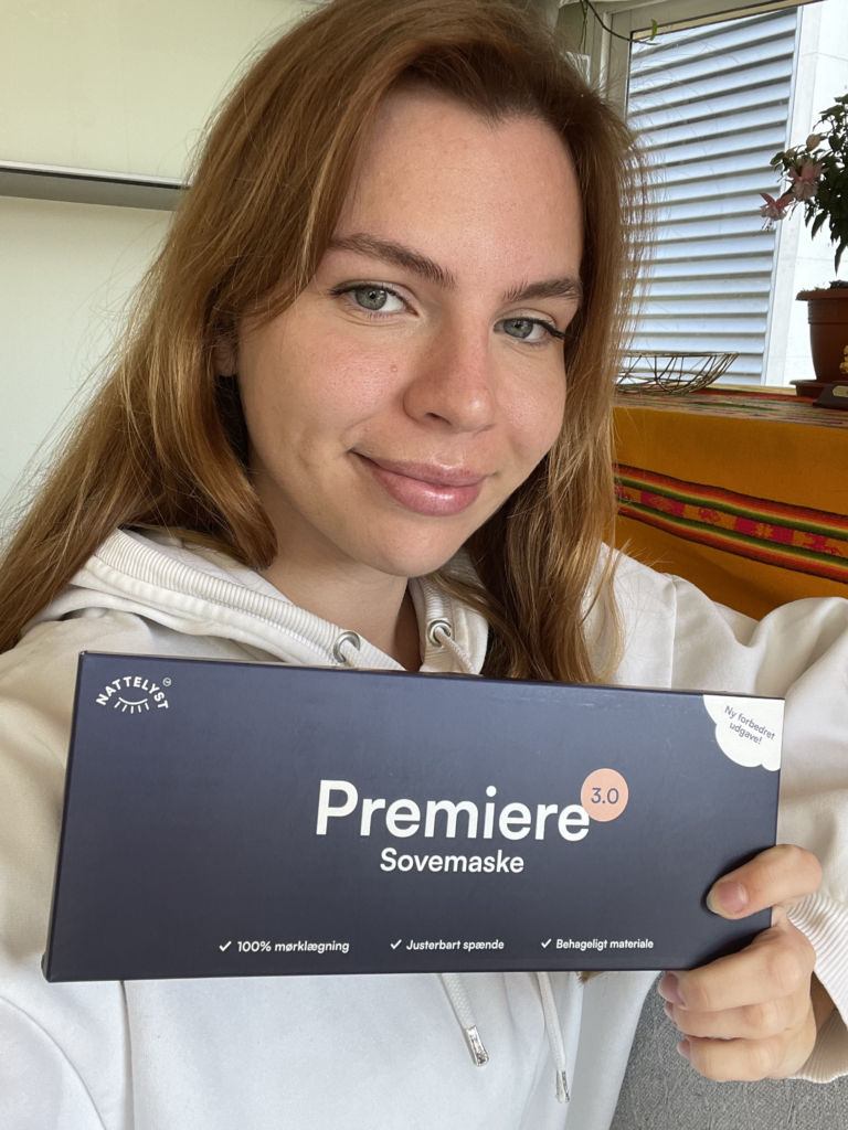 Lächelnde junge Frau in weißer Kapuzenjacke hält Box mit der Aufschrift Premiere Sovemaske, Pflanze im Hintergrund