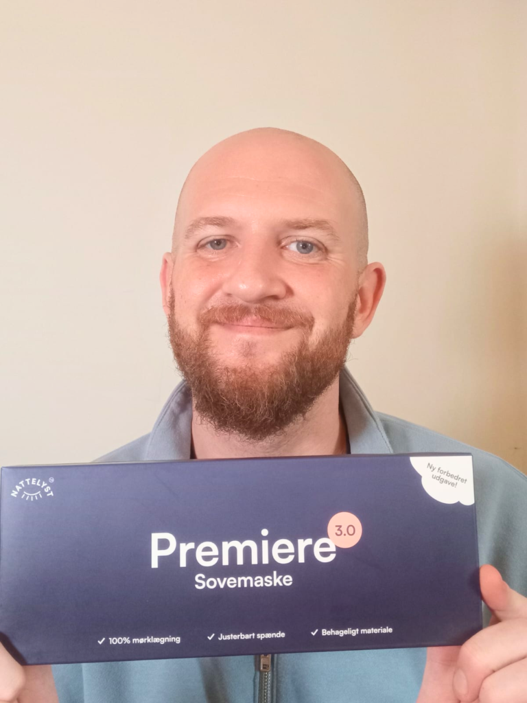 Lächelnder, glatzköpfiger Mann mit Bart hält blaue Verpackung mit Aufschrift Premiere Sovemaske
