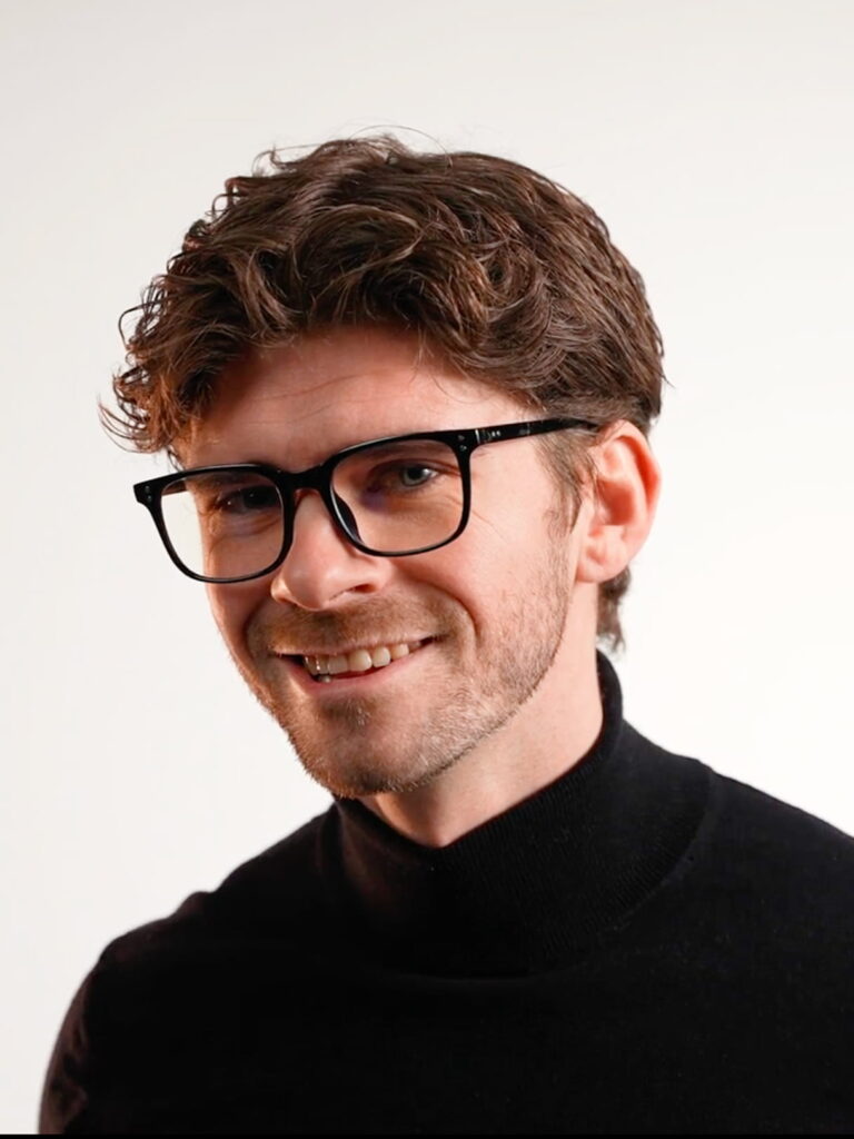 Junger Mann mit dunkler Hornbrille, lockigem braunem Haar, leichter Bartstoppel und schwarzem Rollkragenpullover, lächelnd