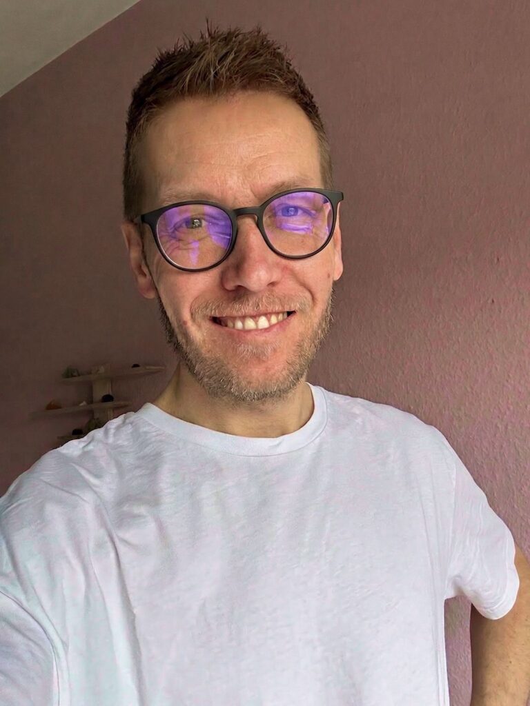 Lächelnder Mann mit runder Brille, in der blaues Licht reflektiert, und weißem T-Shirt vor rosafarbener Wand